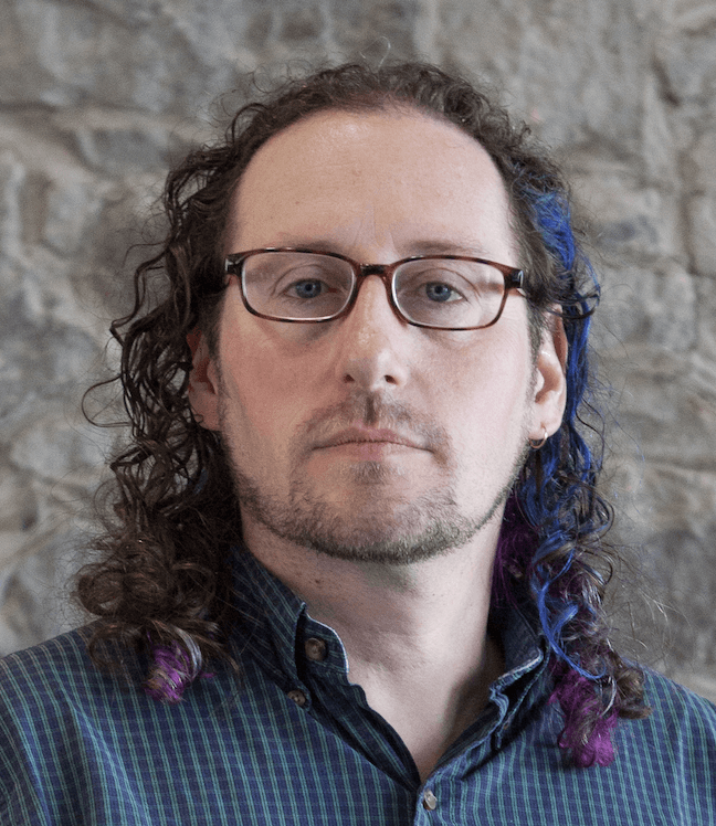 Auteur du portrait. Un homme blanc en 30s/40s avec des lunettes et une touche de bleu dans ses cheveux bruns bouclés.