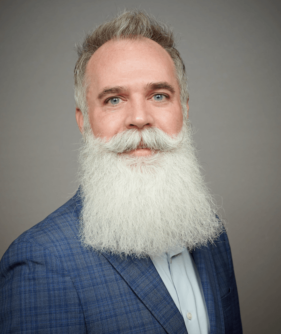 Une photo d’un homme blanc d’âge moyen avec des yeux bleus, des cheveux grisants et une barbe blanche attirant l’attention qui court à mi-chemin sur sa poitrine.