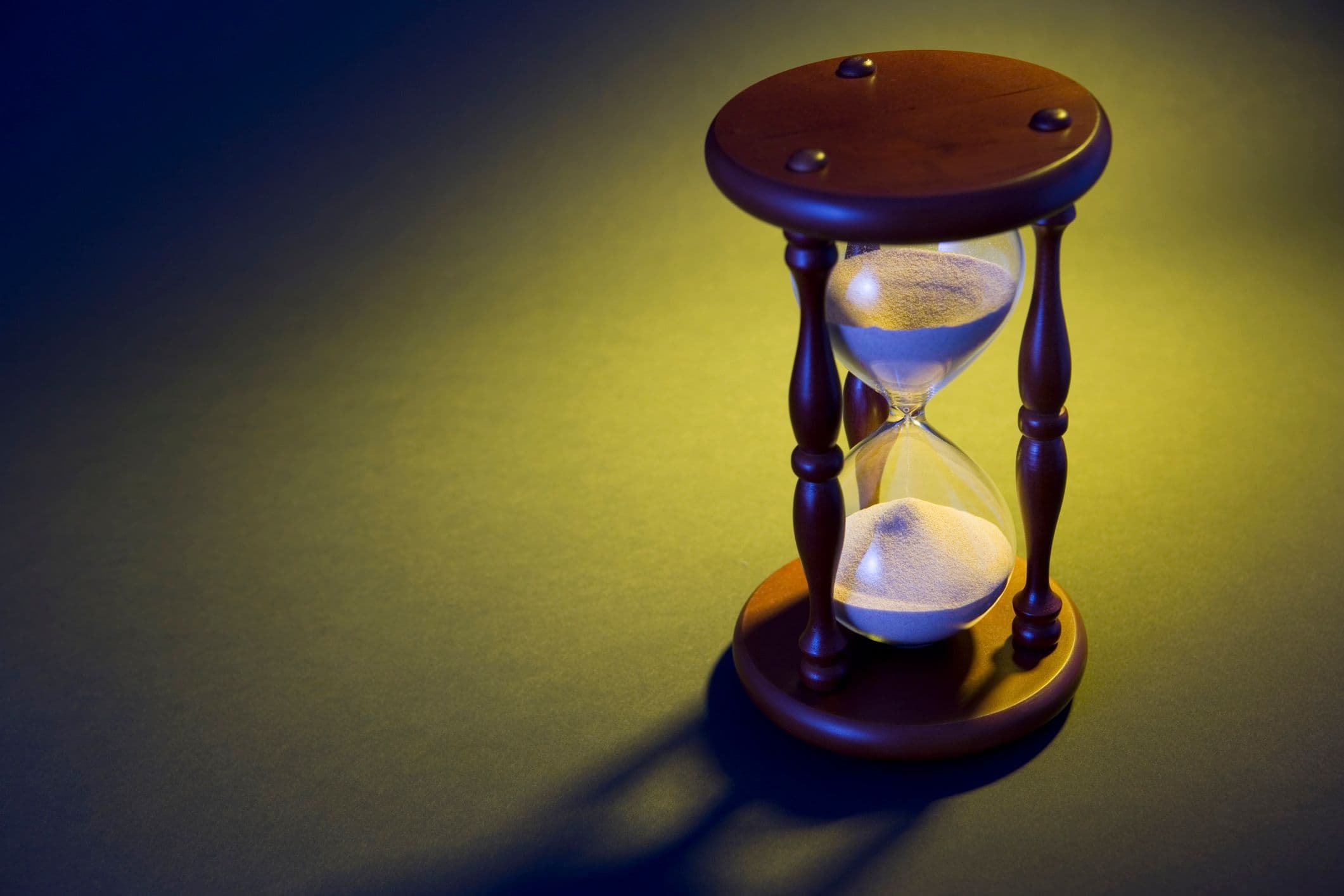 A photo looking down on an hourglass seeping white sand and set against a yellow-ish spotlight.