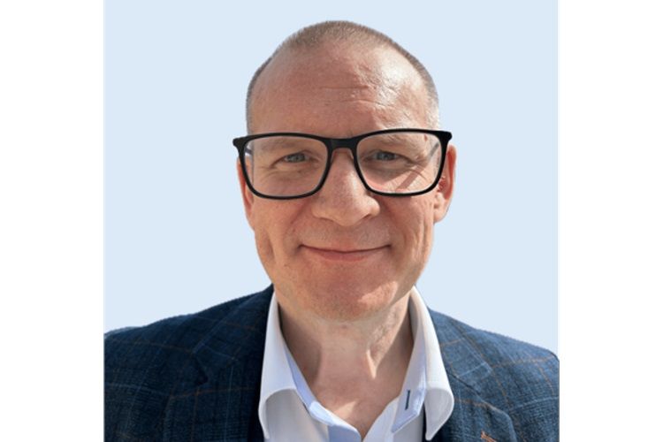 Photo d'un homme blanc aux cheveux coupés très courts et portant des lunettes à monture large, vêtu d'une veste à carreaux grise.