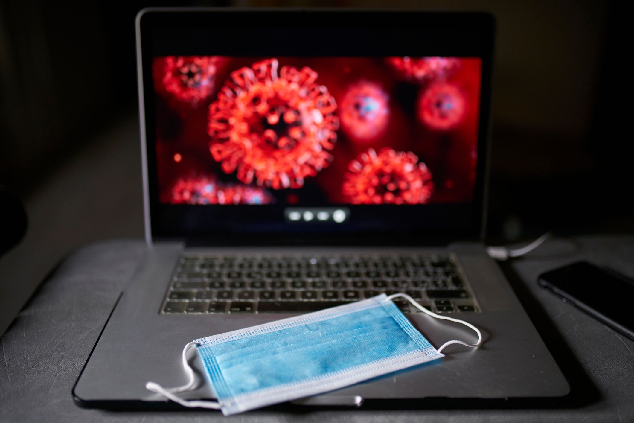 Dans un espace sombre, un masque bleu repose sur le clavier d'un ordinateur portable, tandis que des images microscopiques du virus COVID rouge et hérissé de pointes illuminent l'écran.