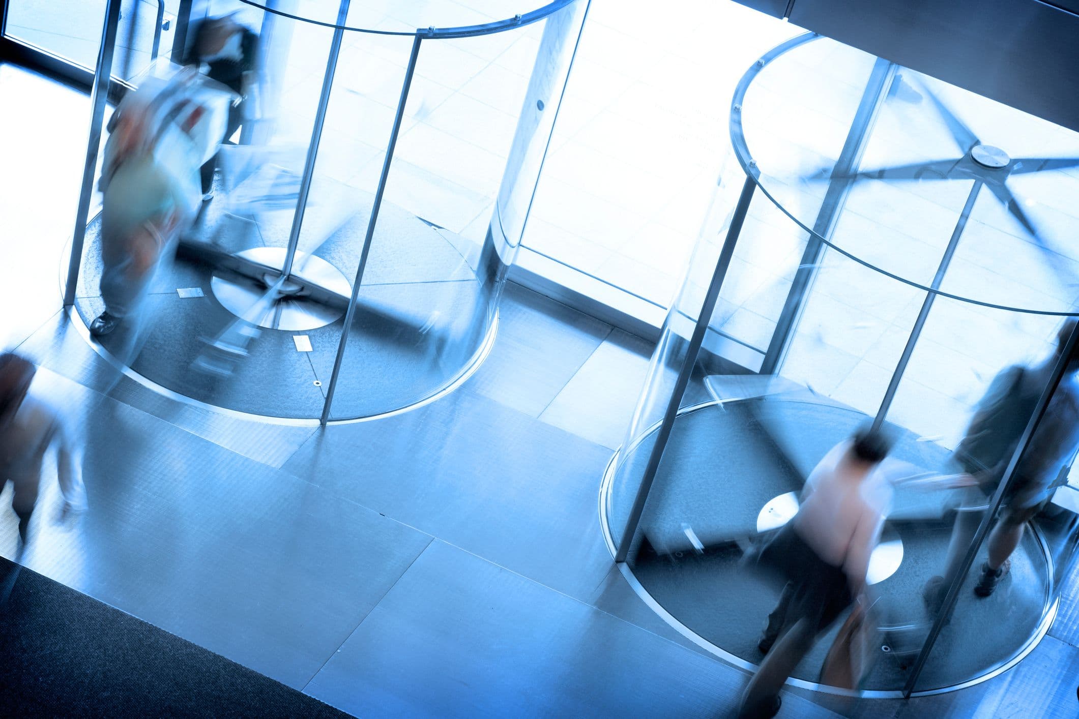 A blurry blue image of people passing through two revolving doors, seen from above and at a diagonal.