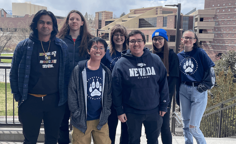 Photo de 7 personnes posant pour une photo de groupe sur une promenade extérieure avec un parc et des bâtiments universitaires en arrière-plan.