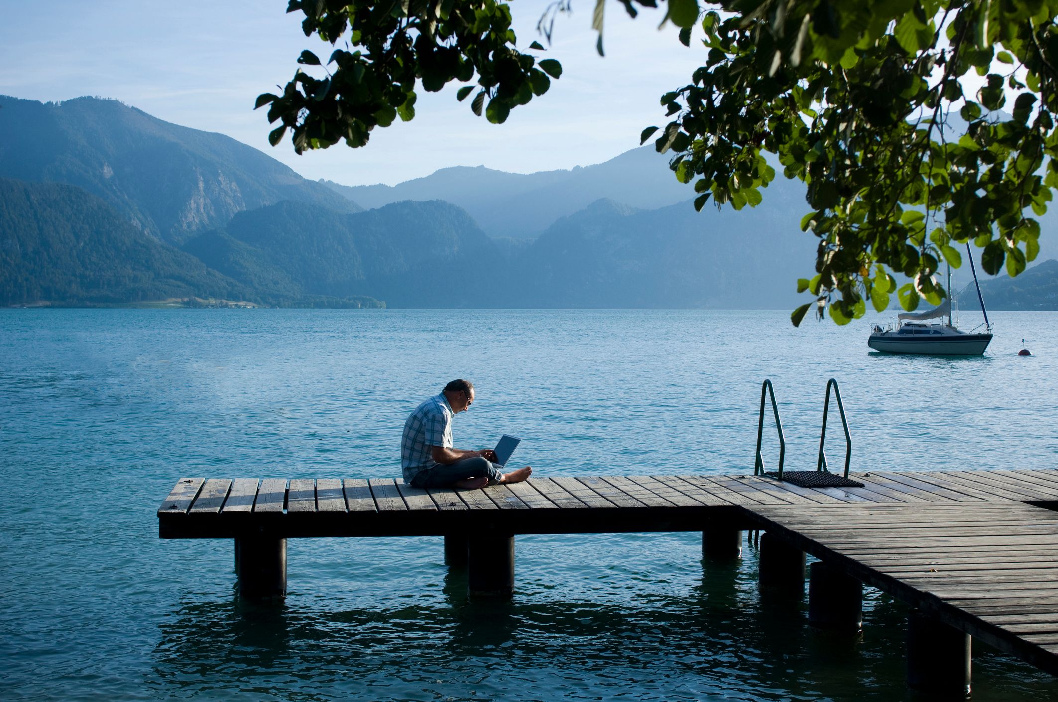 Photo d'un homme assis en tailleur sur un ponton au bord d'un lac, un ordinateur portable sur les genoux.