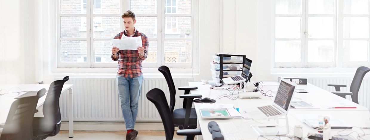 Une personne en chemise en flanelle examine des documents debout près de la fenêtre dans un bureau lumineux aux murs blancs.