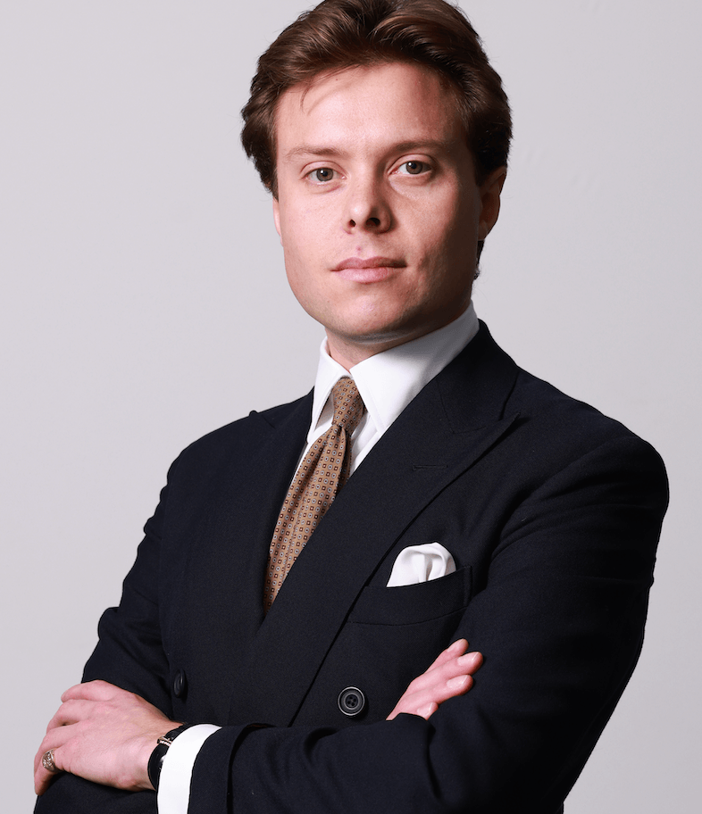 Photo d’un homme blanc 30something aux cheveux bruns, les bras pliés, portant un costume croisé bleu marine impeccable.